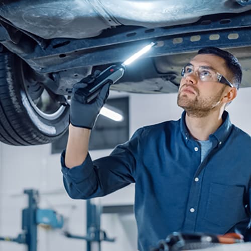 A mechanic wearing safety glasses and gloves inspects the underside of a lifted car in a well-lit automotive workshop, using a handheld flashlight to illuminate the components above him. A mechanic wearing safety glasses and gloves inspects the underside of a lifted car in a well-lit automotive workshop, using a handheld flashlight to illuminate the components above him.